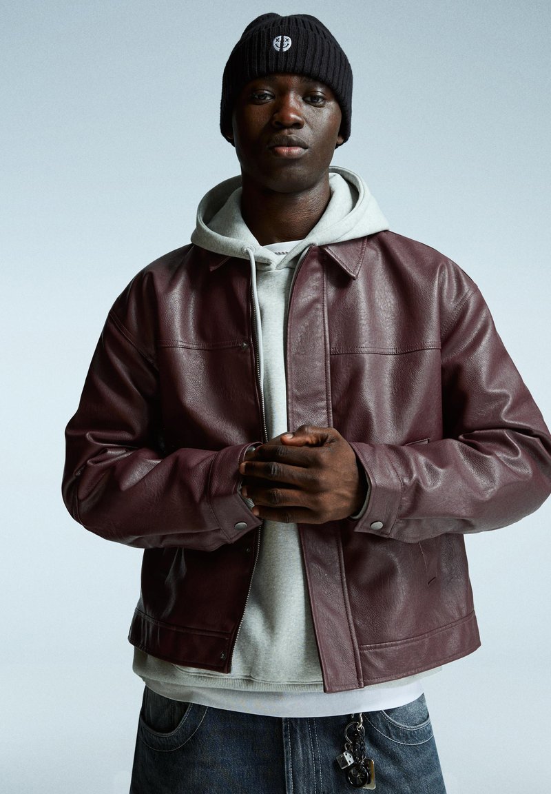 Veste en cuir bourgogne avec un design à col, portée sur un hoodie gris. La personne porte également un bonnet en tricot noir et un jean en denim.