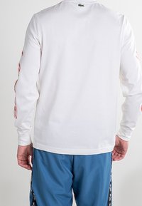 Camicia bianca a maniche lunghe con scollatura rotonda, caratterizzata da un piccolo logo verde sul retro. Abbinata a pantaloni sportivi blu con accenti laterali.