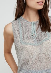Blusa sin mangas de patrón floral ligero, confeccionada en una suave y transparente tela. Presenta un diseño de pinzas y un lazo decorativo en el escote con acentos de encaje.