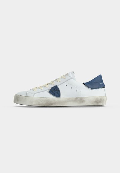 Baskets blanches avec des lacets écrus, un patch bleu au talon, un emblème bleu sur le côté et une semelle volontairement usée et à l’aspect sale sur fond uni.