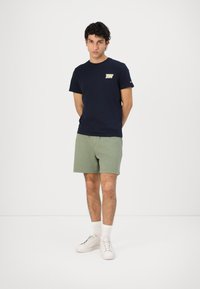 T-shirt en coton bleu marine avec un logo sur la poitrine, associé à un short en coton vert et des baskets blanches, se tenant devant un fond uni.