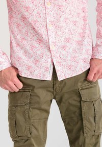 Chemise boutonnée rose clair avec motif floral, portée sur un pantalon cargo vert olive avec poches latérales et un design décontracté.