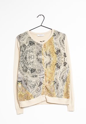 Krémový dlouhý kardigan s černými a zlatými paisley vzory, kulatým výstřihem a zapínáním na knoflíky vpředu, zavěšený na černém ramínku.