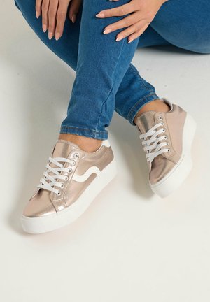 Gambe incrociate con jeans blu, indossando sneakers color oro rosa metallizzato con suole e lacci bianchi, poggiate su una superficie chiara e uniforme.