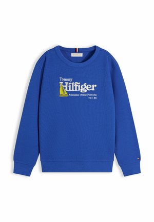 Uśmiechnięty chłopiec stoi z rękami w kieszeniach, ubrany w niebieską bluzę Tommy Hilfiger, luźne niebieskie dżinsy i beżowe trampki na białym tle.