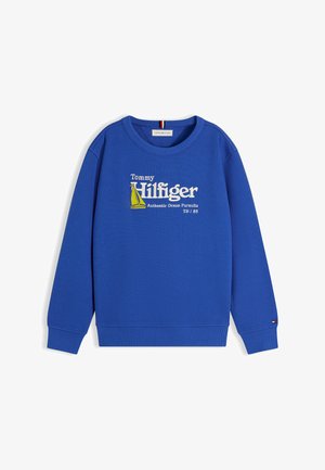 Lachende jongen staand met handen in de zakken, een blauwe Tommy Hilfiger trui dragend, losse blauwe spijkerbroek en beige sneakers op een witte achtergrond.