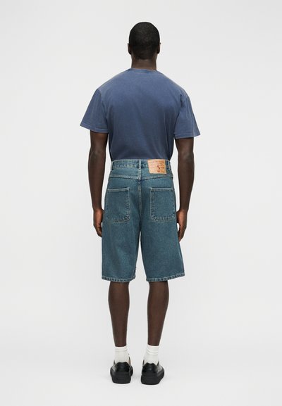 Τζιν σορτς με ψηλή μέση, με δύο πίσω τσέπες. Συνδυάζονται με μπλε-ναυτικό βαμβακερό T-shirt, εμφανίζεται από πίσω. Μαύρα παπούτσια και λευκές κάλτσες.