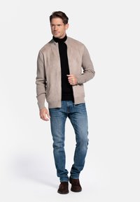 Beige suedine bomberjack met gebreide mouwen, ritssluiting en geribde zoom, gecombineerd met blauwe spijkerbroek en bruine suède laarzen.