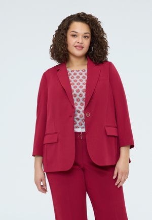 Femme aux cheveux bouclés portant une veste et un pantalon de costume rouge sur un haut à motifs, debout devant un fond clair uni.