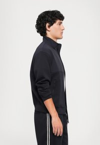 Ung mann i svart zip-up jakke og matchende bukser med hvite striper står i profil mot en lys bakgrunn.