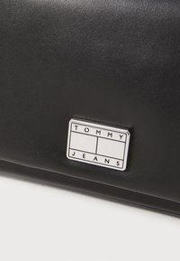 Svart syntetisk lær clutch med en glatt tekstur, med en sølvfarget rektangulær logo-plakett merket "TOMMY JEANS."