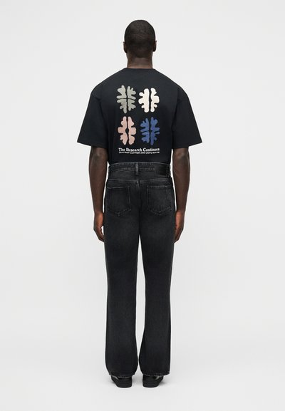 Άνδρας που φοράει μαύρο μπλουζάκι με τέσσερα πολύχρωμα αφηρημένα σχήματα και κείμενο, συνδυασμένο με μαύρο τζιν φαρδιάς γραμμής και μαύρα παπούτσια, γυρισμένος προς τα πίσω.