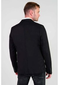 Zwarte blazer met gestructureerde schouders, enkele split en knoopmanchetten. Stof lijkt glad en op maat, gecombineerd met een donkere jeans.