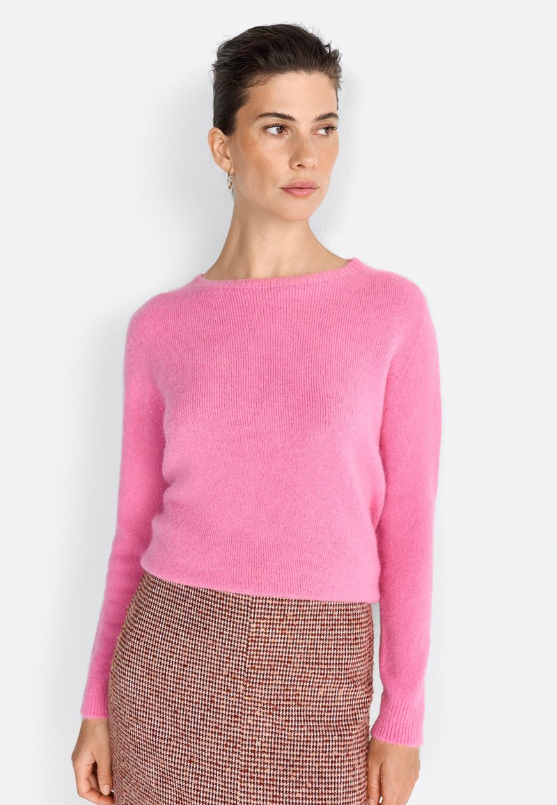 Pulover roz, pufos, din tricot, cu gât rotund și mâneci lungi; asortat cu o fustă cu carouri maro, dintr-un material texturat.