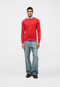 Maglione rosso lavorato a maglia, maniche lunghe, collo rotondo; indossato sopra una camicia a colletto blu chiaro. Abbinato a jeans blu chiaro e scarpe marroni.