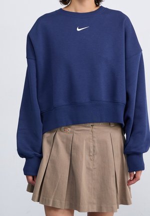 Osoba nosząca granatową bluzę Nike i beżową plisowaną spódnicę z małym logo Dickies na pasku.