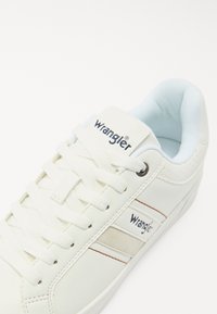 Baskets en cuir blanc avec des accents en textile, présentant des rayures brunes et une langue de chaussure marquée. Les lacets sont blancs et l'intérieur est bleu clair.