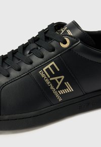Svarte lær sneakers med gullmerke, svarte snørebånd og gummisåle. Siden har en logo med "EA" og teksten "EMPORIO ARMANI".