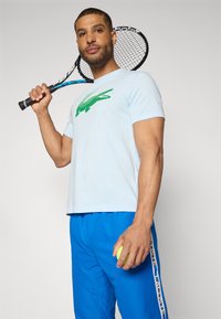Šviesiai mėlyna medvilninė marškinė su žalia krokodilo grafika, derinama su ryškiai mėlynomis sportinėmis kelnėmis, turinčiomis baltą "LACOSTE" juostą, laikant teniso raketę ir kamuoliuką.