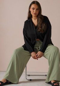 Svart knapp-up skjorta, avslappnad passform; ljusgröna vida byxor, texturerat tyg; svarta sandaler med en liten guldkant; minimalistisk miljö.