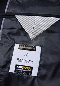 Schwarzer Satin-Stoff mit einer diagonalen grauen gestreiften Taschenfutter. An den Etiketten sind "Endurance", "Officina by Marlane" und "CORDURA" sichtbar.