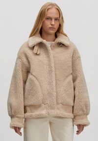 Beige sheepskin jas met een grote kraag, zijzakken en leren riemaccenten. Boxy design met een zachte textuur en geribbelde manchetten.