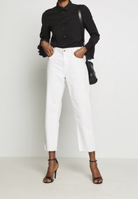 Camicia nera con bottoni e polsini plissettati, abbinata a jeans bianchi cropped con orli grezzi. I sandali neri con cinturini completano l'outfit.
