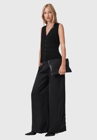 Czarny bezrękawnik z dekoltem w kształcie litery V oraz guzikami, w połączeniu z szerokimi czarnymi spodniami. Modelka trzyma czarną torebkę kopertówkę, o gładkiej fakturze materiału.