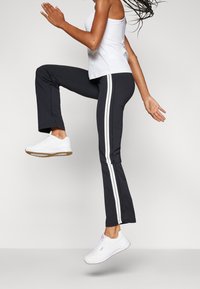 Černé sportovní kalhoty s bílými pruhy na bocích, doplněné bílým tílkem a bílými teniskami. Materiál vypadá hladký; kalhoty mají rozšířený spodní lem.