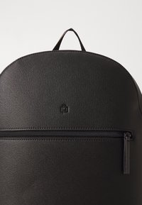 Černý batoh s texturovaným povrchem, zaobleným tvarem, přední kapsou na zip a malým logem. Horní rukojeť pro snadné nošení.