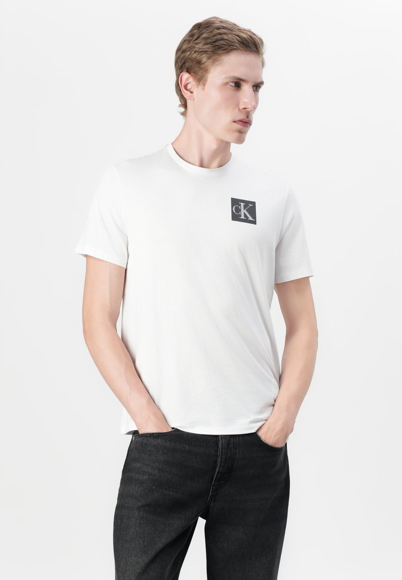 T-shirt en coton blanc à manches courtes, avec un patch carré noir arborant le logo 'cK' sur la poitrine gauche. Le modèle porte un jean noir.