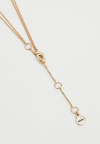 Collier en ton or avec deux chaînes, arborant des maillons ronds et une étiquette gravée "DKNY". La texture est lisse, avec une apparence délicate.