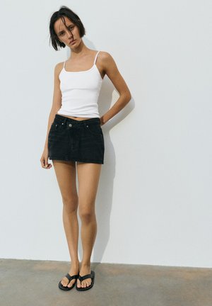 Žena u bijelom topu, crnoj traper mini suknji i crnim sandalama stoji naslonjena na bijeli zid s jednom rukom iza leđa.