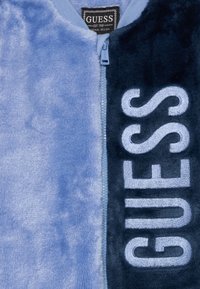 Kailinys džemperis su mėlynomis ir tamsiai mėlynomis dalimis, su siuvinėtu "GUESS" logotipu, pilnu užtrauktuku ir švelnia tekstūra.