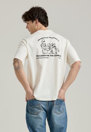 Cream-colored cotton t-shirt with short sleeves, featuring a black graphic design and text on the back. Worn with blue jeans.