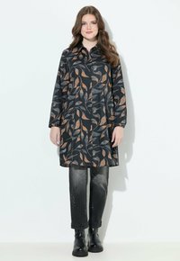 Femme avec de longs cheveux bruns portant une tunique noire à boutons avec des motifs de feuilles orange et grises, un jean noir délavé et des bottes noires.