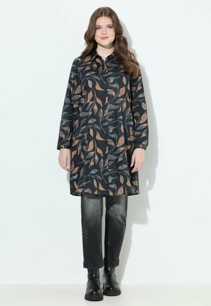 Femme avec de longs cheveux bruns portant une tunique noire à boutons avec des motifs de feuilles orange et grises, un jean noir délavé et des bottes noires.