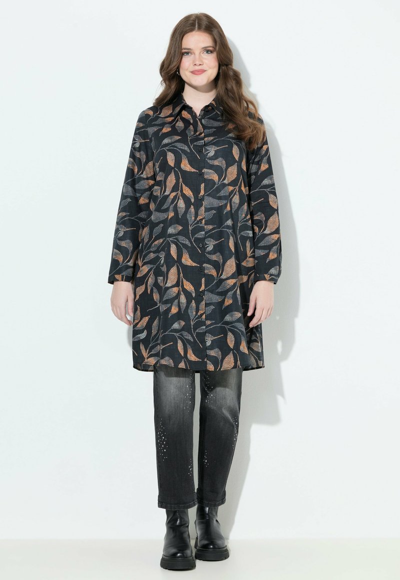 Femme avec de longs cheveux bruns portant une tunique noire à boutons avec des motifs de feuilles orange et grises, un jean noir délavé et des bottes noires.