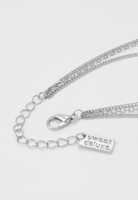 Sidabrinė grandinėlė su krabų užsegimu, turinti susipynusias sruogas ir stačiakampį tagą, ant kurio įspaustas "sweet deluxe."