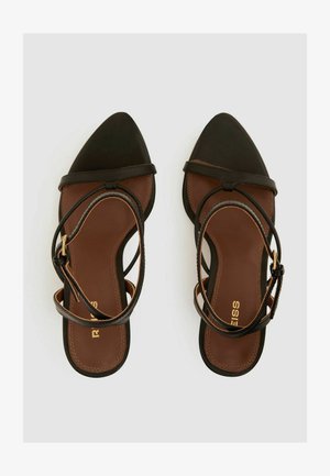 Fekete pántos szandál, hegyes orral, barna talppal és vékony bokapántokkal. Simas bőr textúrával és arany színű kiegészítőkkel rendelkezik.