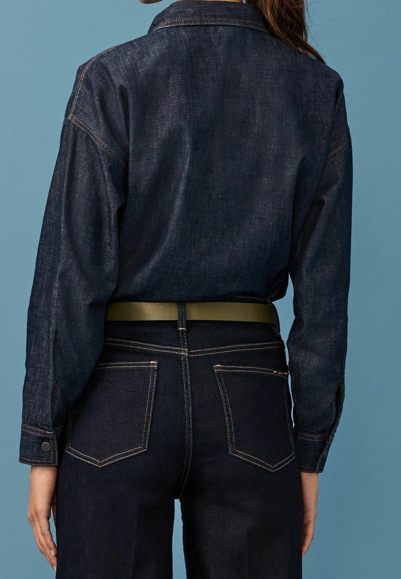 Personne portant une chemise en denim foncé rentrée dans un pantalon en denim foncé assorti, avec une ceinture vert olive, vue de dos sur un fond bleu.