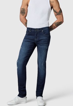 Mann iført mørkeblå slitte jeans, hvit tanktop og hvite joggesko, står med hendene bak ryggen på lys grå bakgrunn.