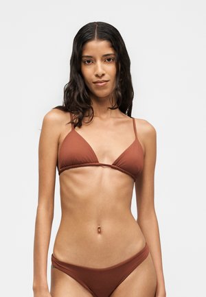 Vrouw met lang donker haar, gekleed in een bruin driehoek-bikinitopje en bijpassende broekjes, staand tegen een effen lichte achtergrond.