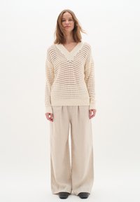 Beige gestrickter Pullover mit V-Ausschnitt und Lochmuster, kombiniert mit lockeren hellbeigen Leinenhosen und schwarzen Schuhen.