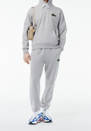 Persona che indossa una tuta Lacoste grigio chiaro con felpa con cappuccio e pantaloni della tuta abbinati, porta una borsa a tracolla beige e indossa sneakers bianche e blu.