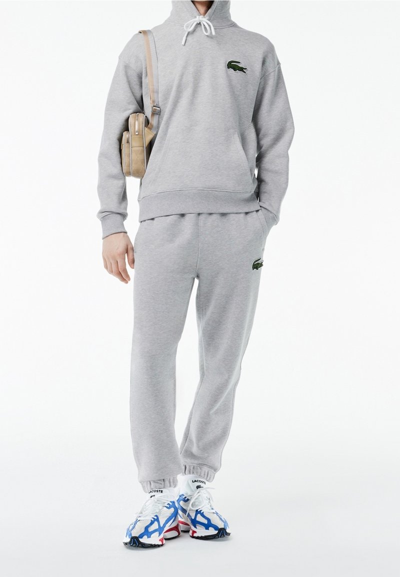 Personne portant un survêtement Lacoste gris clair avec un sweat à capuche et un pantalon de jogging assortis, portant un sac à bandoulière beige et des baskets blanches et bleues.