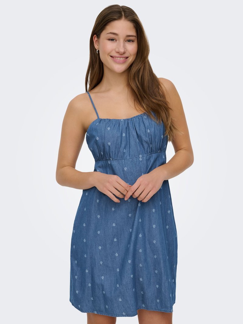 Jeune femme souriante, portant une robe bleue sans manches avec de petits motifs clairs, debout devant un fond clair uni.