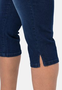 Tmavě modré džíny capri s přiléhavým střihem. Disponují bočními rozparky na lemech a hladkou texturou.