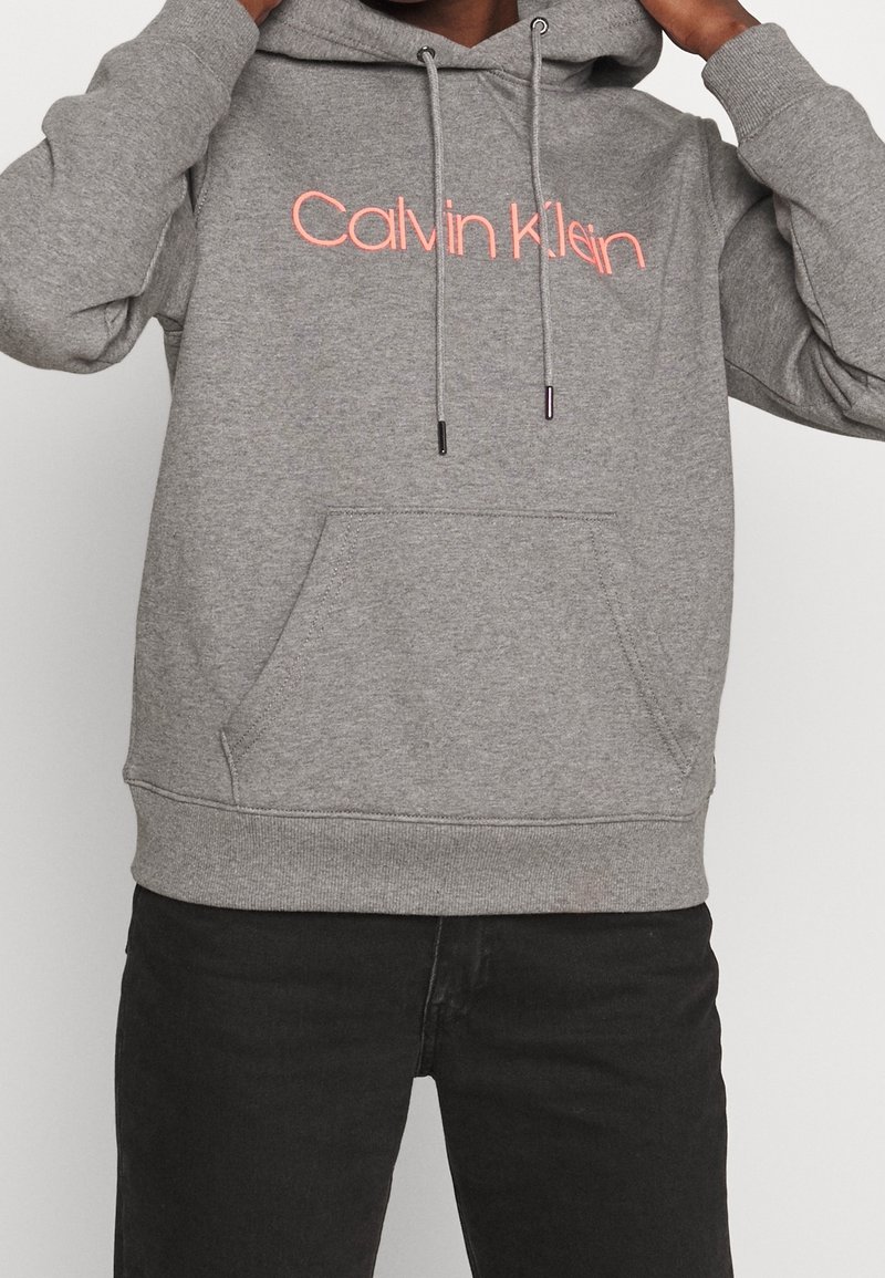 Felpa grigia Calvin Klein con tasca a marsupio, cordini regolabili e logo ricamato rosa sul petto. Tessuto morbido.
