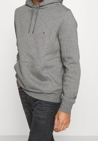 Sudadera gris de mezcla de algodón suave, con un bolsillo tipo canguro en el frente, cordones de metal y un pequeño logo en el pecho.